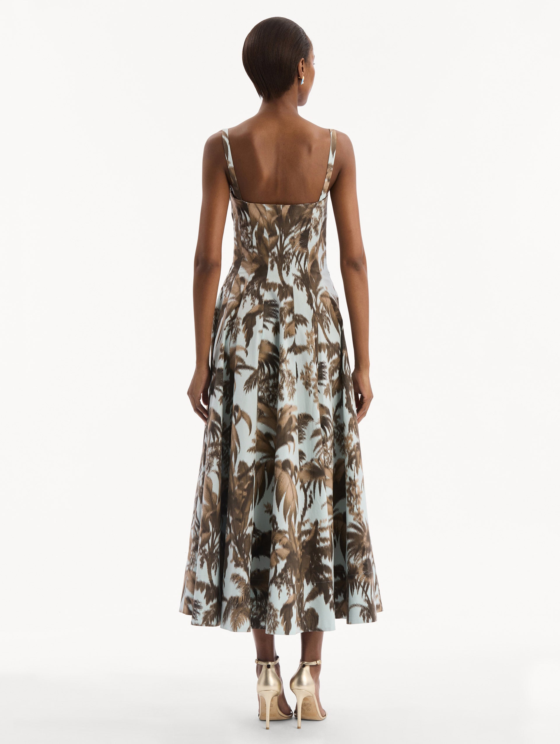 The model is wearing the Chiné Palm Leaves Cotton Poplin Dress in light blue printed with brown palm leaves. Featuring a subtle sweetheart neckline with tank straps, a softly structured bodice, and flowing midi skirt. Back View.
