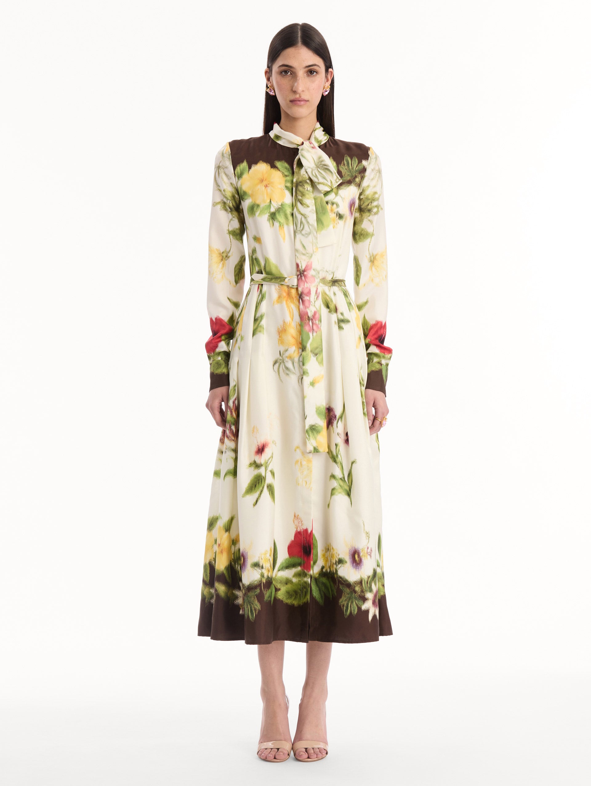 The model is wearing the Chiné Dominican Mixed-Floral Silk Twill Dress. Featuring a vibrant array of Dominican florals, tie neckline, belt at waist, long sleeves, and a midi length. Front View.
