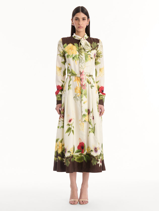 The model is wearing the Chiné Dominican Mixed-Floral Silk Twill Dress. Featuring a vibrant array of Dominican florals, tie neckline, belt at waist, long sleeves, and a midi length. Front View.