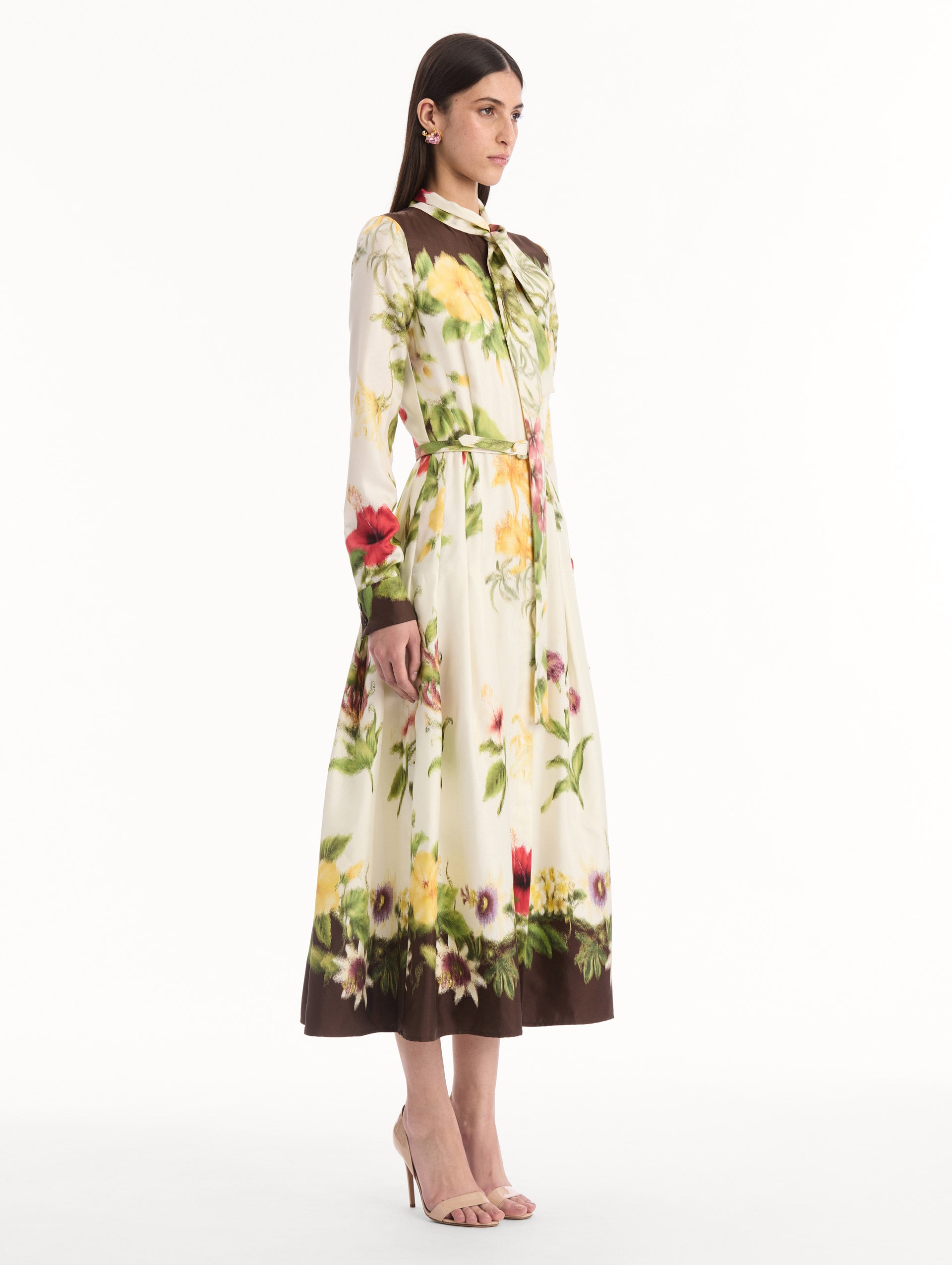 The model is wearing the Chiné Dominican Mixed-Floral Silk Twill Dress. Featuring a vibrant array of Dominican florals, tie neckline, belt at waist, long sleeves, and a midi length. Side View.