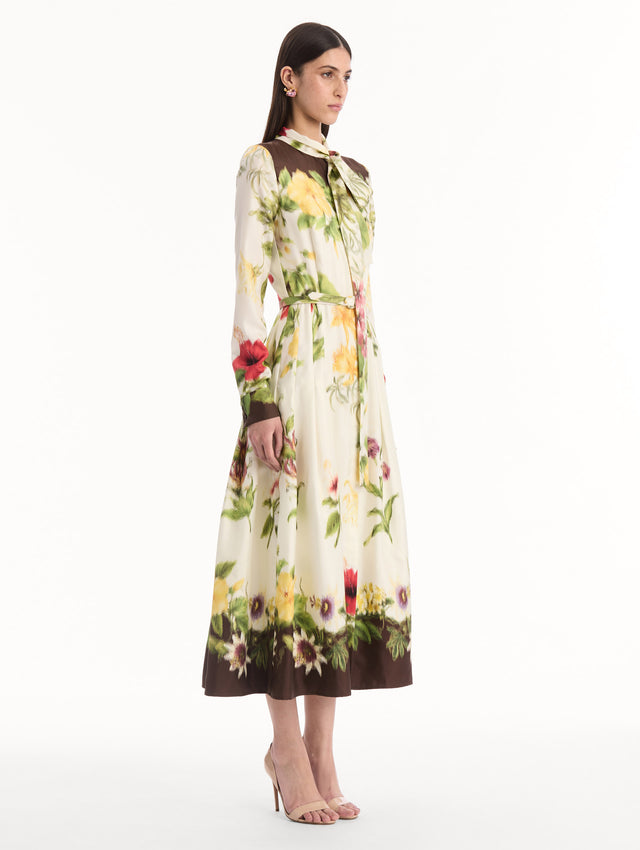 The model is wearing the Chiné Dominican Mixed-Floral Silk Twill Dress. Featuring a vibrant array of Dominican florals, tie neckline, belt at waist, long sleeves, and a midi length. Side View.