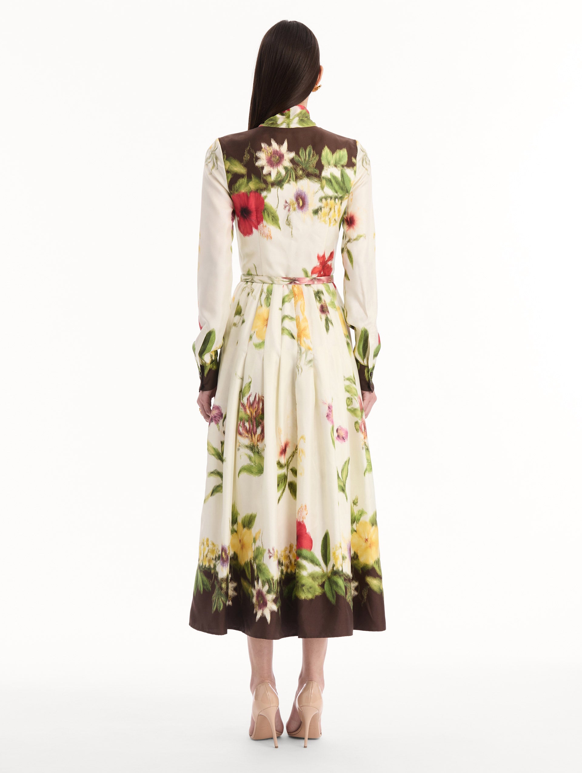 The model is wearing the Chiné Dominican Mixed-Floral Silk Twill Dress. Featuring a vibrant array of Dominican florals, tie neckline, belt at waist, long sleeves, and a midi length. Back View.