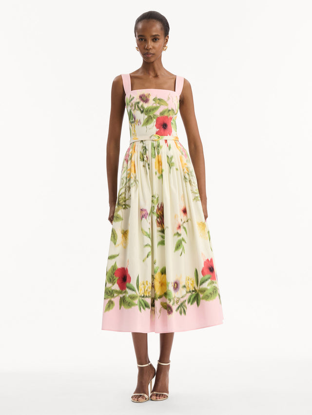 The model is wearing the Dominican Mixed-Floral Cotton Poplin Dress in ivory printed with an array of Dominican florals and light pink at the trims. Featuring a square neckline with tank straps, belt at waist, and pleated midi skirt. Front View.