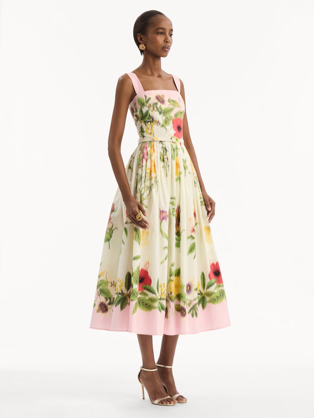 The model is wearing the Dominican Mixed-Floral Cotton Poplin Dress in ivory printed with an array of Dominican florals and light pink at the trims. Featuring a square neckline with tank straps, belt at waist, and pleated midi skirt. Side View.