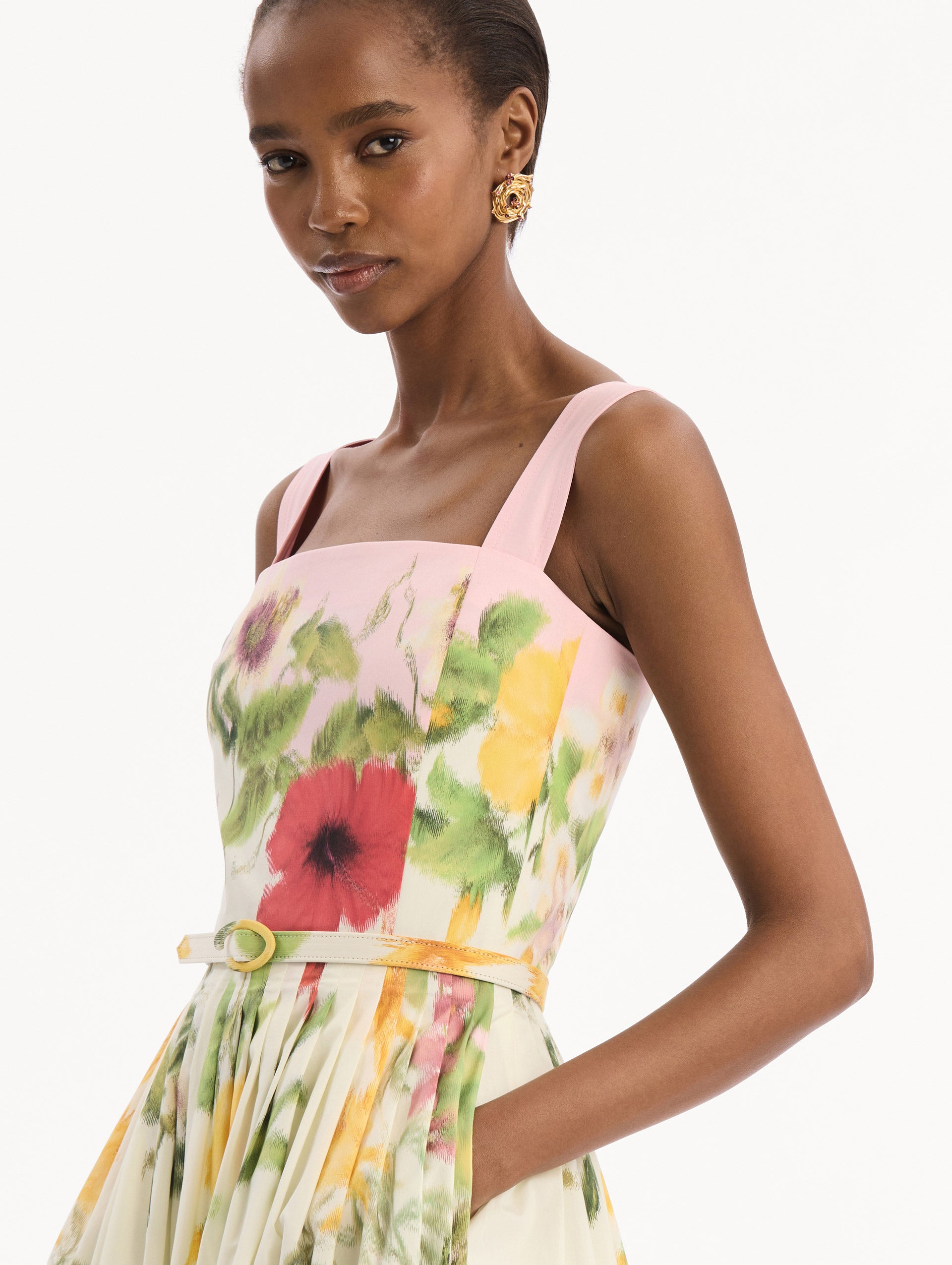 The model is wearing the Dominican Mixed-Floral Cotton Poplin Dress in ivory printed with an array of Dominican florals and light pink at the trims. Featuring a square neckline with tank straps, belt at waist, and pleated midi skirt. Detail View.