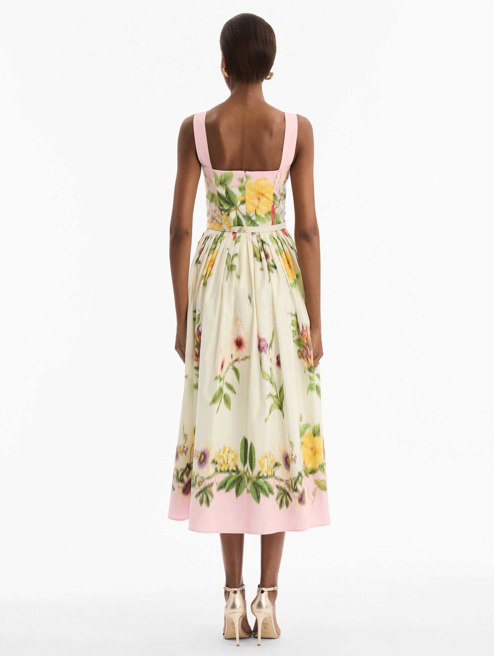 The model is wearing the Dominican Mixed-Floral Cotton Poplin Dress in ivory printed with an array of Dominican florals and light pink at the trims. Featuring a square neckline with tank straps, belt at waist, and pleated midi skirt. Back View.