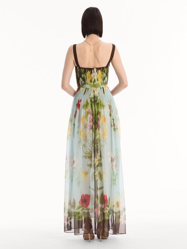 The model is wearing the Dominican Mixed-Floral Silk Chiffon Gown in light blue printed with an array of Dominican florals and brown at the trims. Featuring a pleated bodice, tank straps, tie at waist, and long, flowing skirt. Back View.