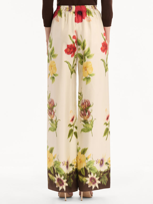 The model is wearing the Dominican Mixed-Floral Wide Leg Pant in ivory printed with an array of Dominican florals. Featuring wide legs and an elastic waistband. Back View.