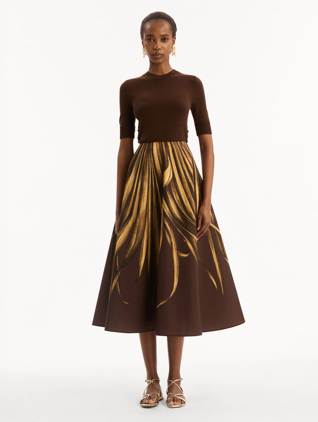 The model is wearing the Palm Leaves Faille Skirt in brown printed with gold palm leaves. Featuring a voluminous, structured silhouette and midi length. Front View.