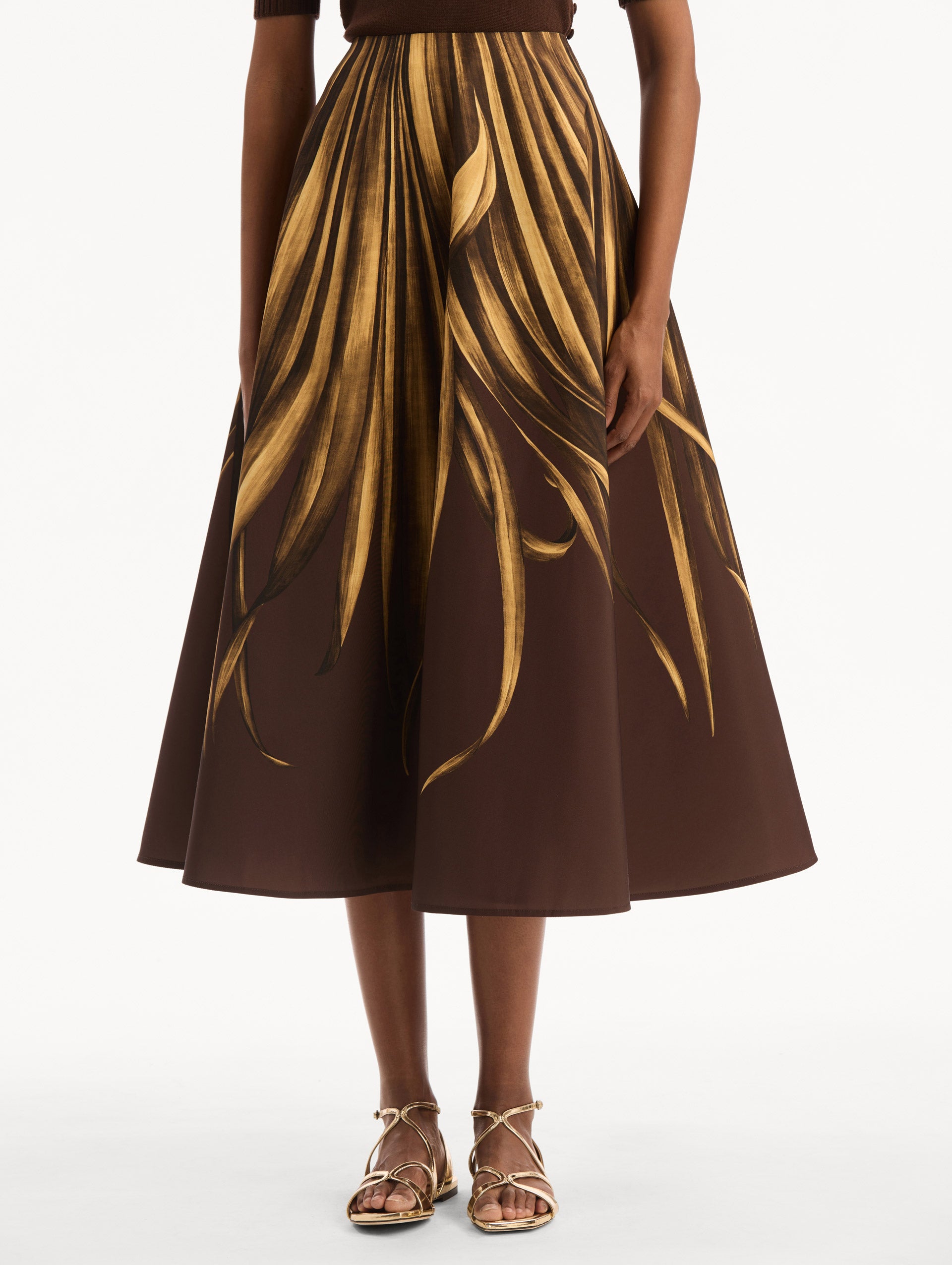 The model is wearing the Palm Leaves Faille Skirt in brown printed with gold palm leaves. Featuring a voluminous, structured silhouette and midi length. Front View.