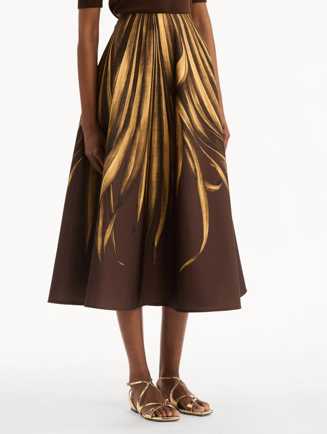 The model is wearing the Palm Leaves Faille Skirt in brown printed with gold palm leaves. Featuring a voluminous, structured silhouette and midi length. Side View.