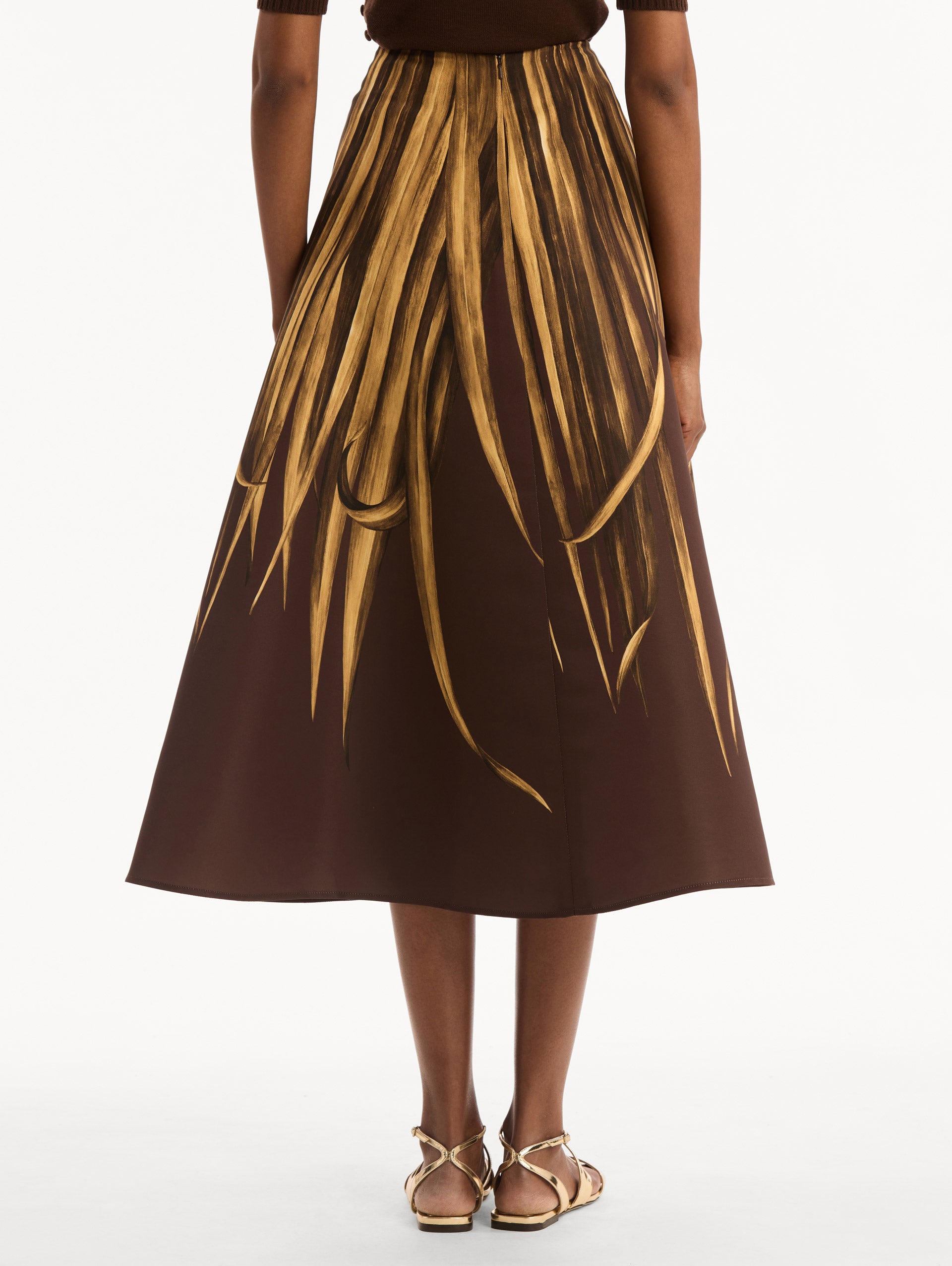 The model is wearing the Palm Leaves Faille Skirt in brown printed with gold palm leaves. Featuring a voluminous, structured silhouette and midi length. Back View.