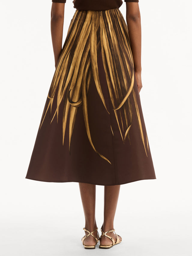 The model is wearing the Palm Leaves Faille Skirt in brown printed with gold palm leaves. Featuring a voluminous, structured silhouette and midi length. Back View.