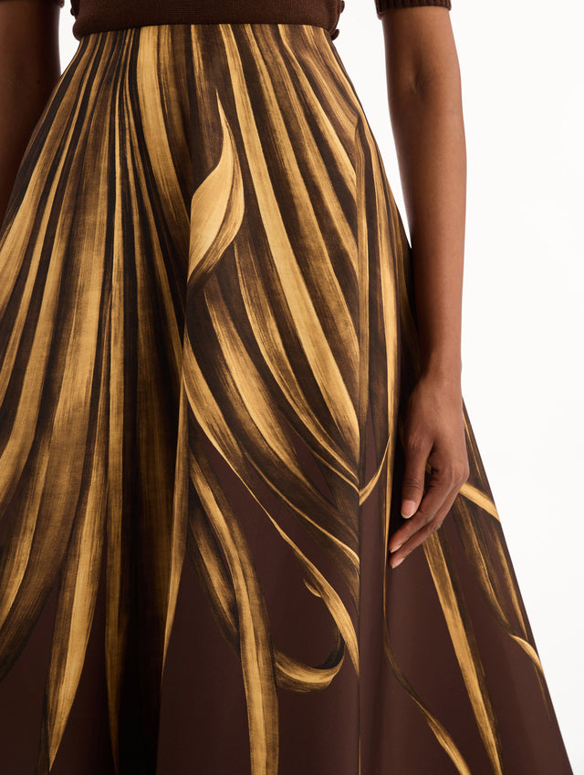 The model is wearing the Palm Leaves Faille Skirt in brown printed with gold palm leaves. Featuring a voluminous, structured silhouette and midi length. Detail View.
