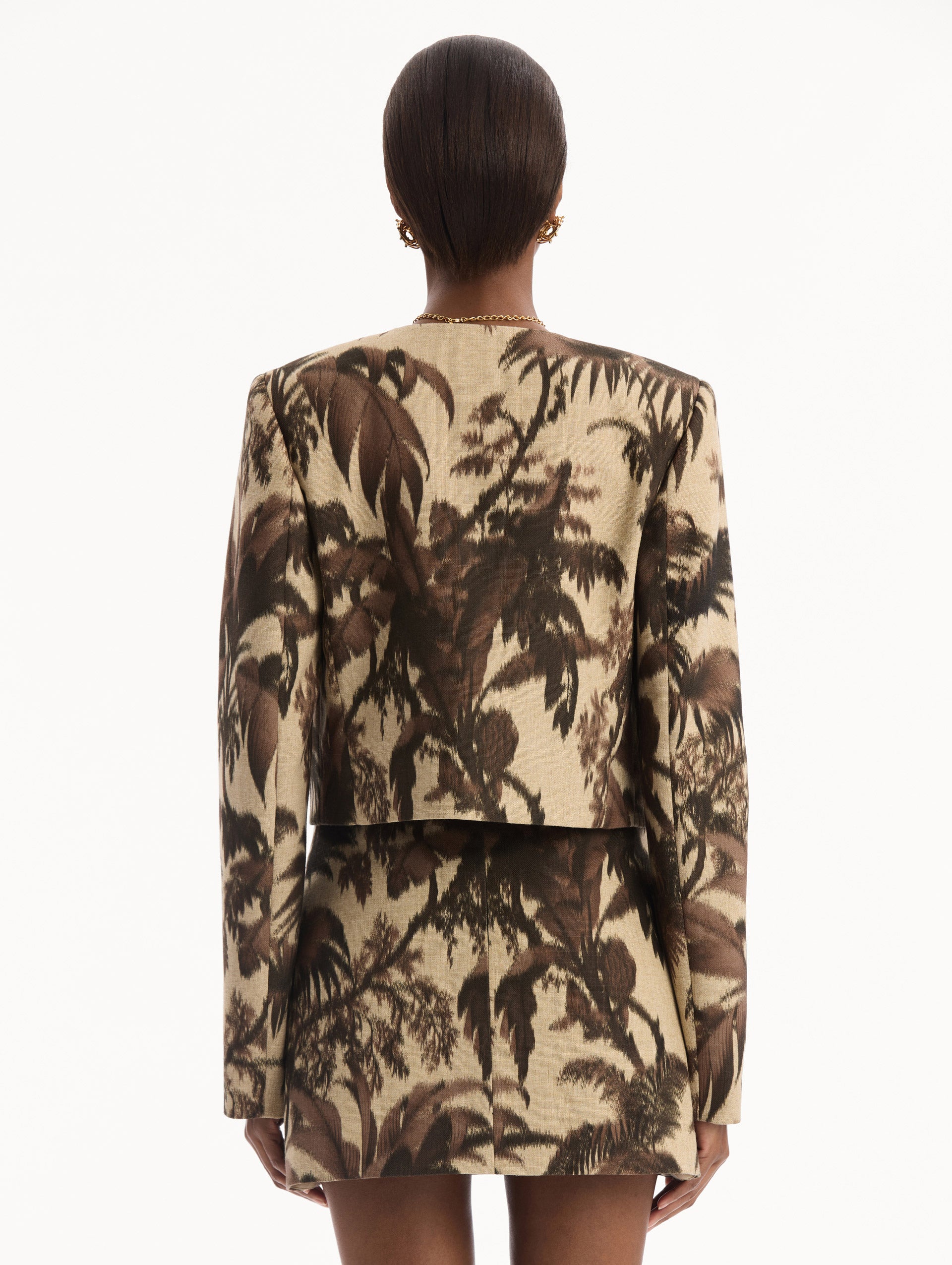 The model is wearing the Chiné Palm Leaves Jacket printed with brown palm leaves. Featuring long sleeves, front pockets with a button closure, and a tailored silhouette. Back View.