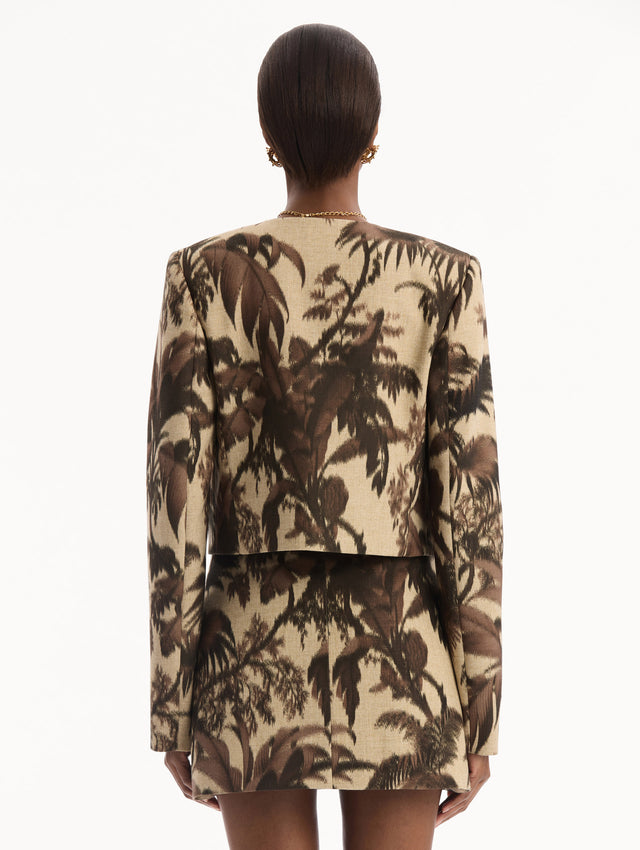 The model is wearing the Chiné Palm Leaves Jacket printed with brown palm leaves. Featuring long sleeves, front pockets with a button closure, and a tailored silhouette. Back View.