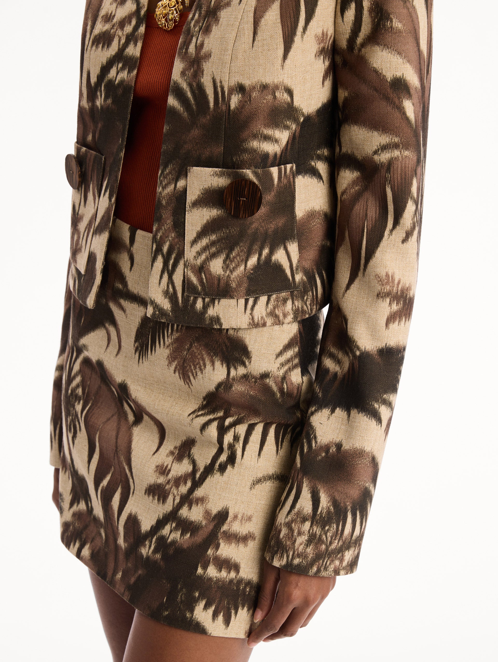 The model is wearing the Chiné Palm Leaves Jacket printed with brown palm leaves. Featuring long sleeves, front pockets with a button closure, and a tailored silhouette. Detail View.