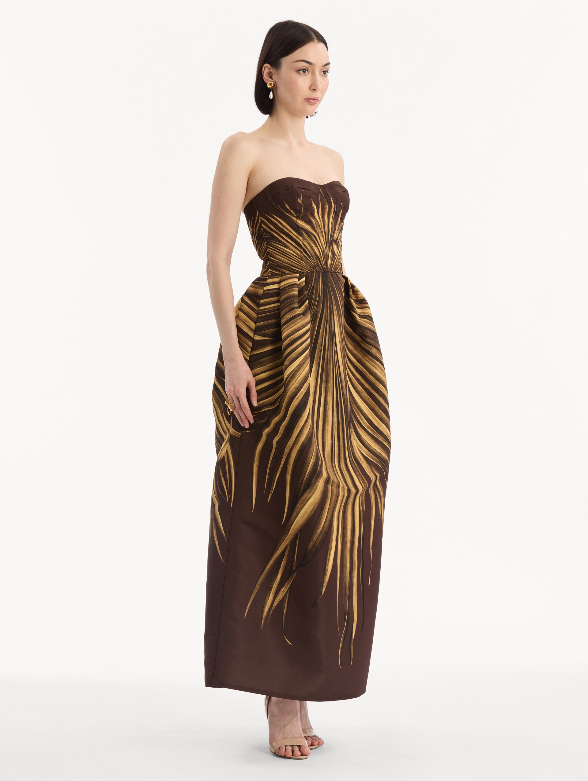 The model is wearing the Palm Leaves Faille Gown in brown printed with gold palm leaves. Featuring a strapless neckline, sculpted bodice, and long skirt. Side View.