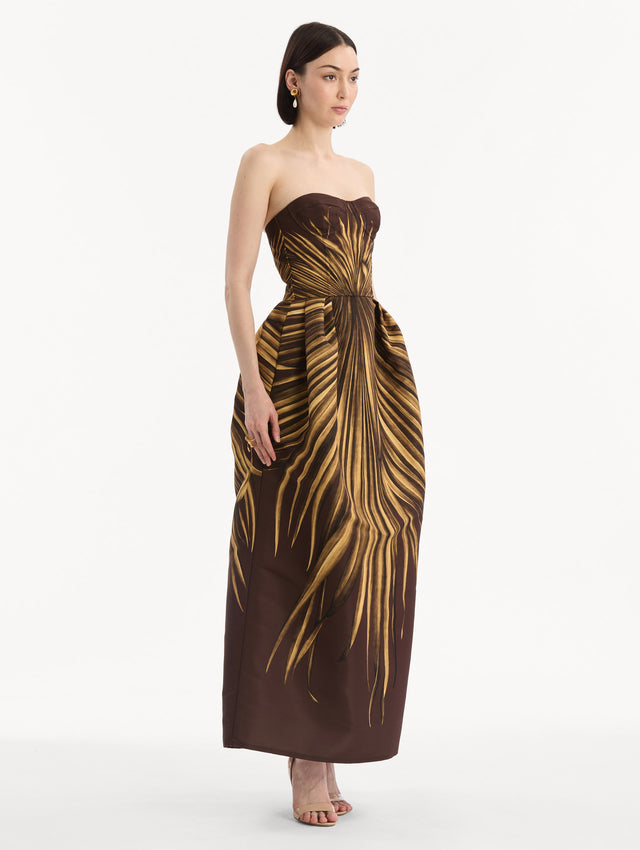 The model is wearing the Palm Leaves Faille Gown in brown printed with gold palm leaves. Featuring a strapless neckline, sculpted bodice, and long skirt. Side View.
