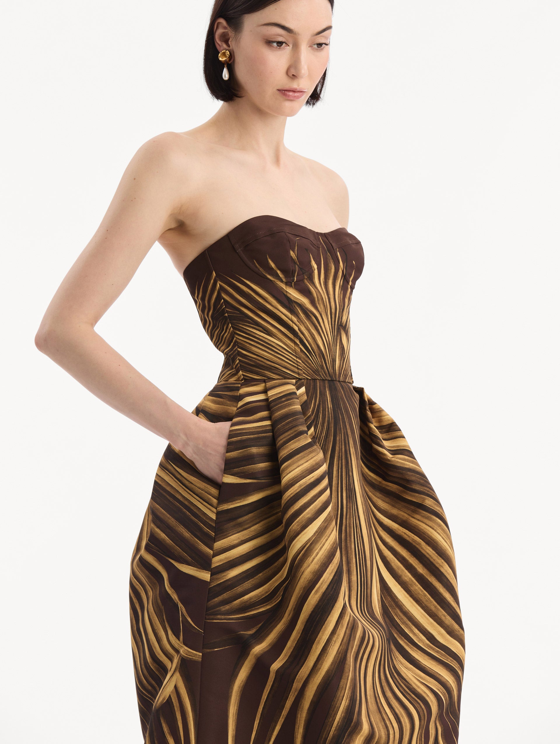 The model is wearing the Palm Leaves Faille Gown in brown printed with gold palm leaves. Featuring a strapless neckline, sculpted bodice, and long skirt. Detail View.