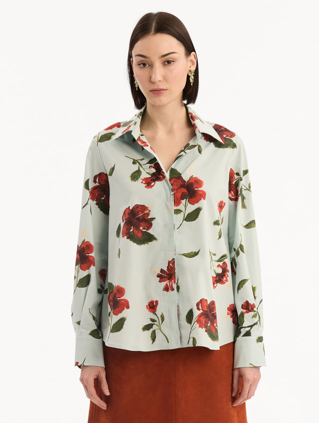 The model is wearing the Hibiscus Cotton Poplin Blouse in a muted blue color printed with red hibiscus florals. Featuring a classic collar, button-front closure, long sleeves, and relaxed silhouette. Front View.