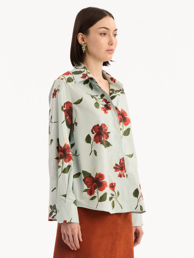 The model is wearing the Hibiscus Cotton Poplin Blouse in a muted blue color printed with red hibiscus florals. Featuring a classic collar, button-front closure, long sleeves, and relaxed silhouette. Side View.