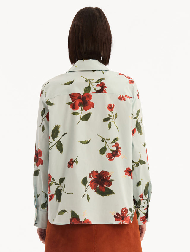 The model is wearing the Hibiscus Cotton Poplin Blouse in a muted blue color printed with red hibiscus florals. Featuring a classic collar, button-front closure, long sleeves, and relaxed silhouette. Back View.