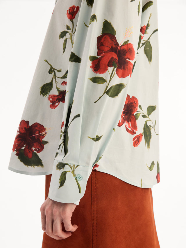The model is wearing the Hibiscus Cotton Poplin Blouse in a muted blue color printed with red hibiscus florals. Featuring a classic collar, button-front closure, long sleeves, and relaxed silhouette. Detail View.