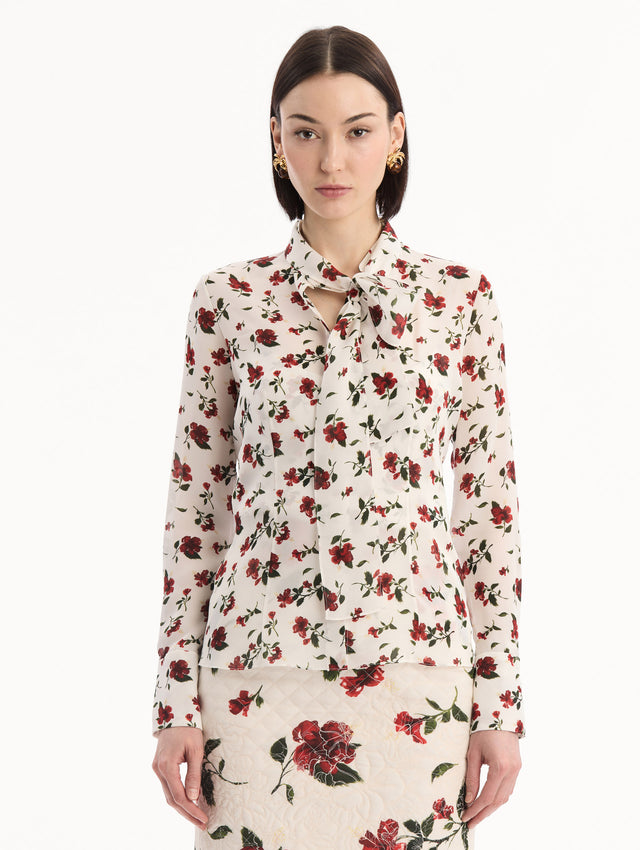 The model is wearing the Hibiscus Silk Chiffon Blouse in white printed with red hibiscus florals. Featuring a tie at neckline, long sleeves, and tailored silhouette. Front View.