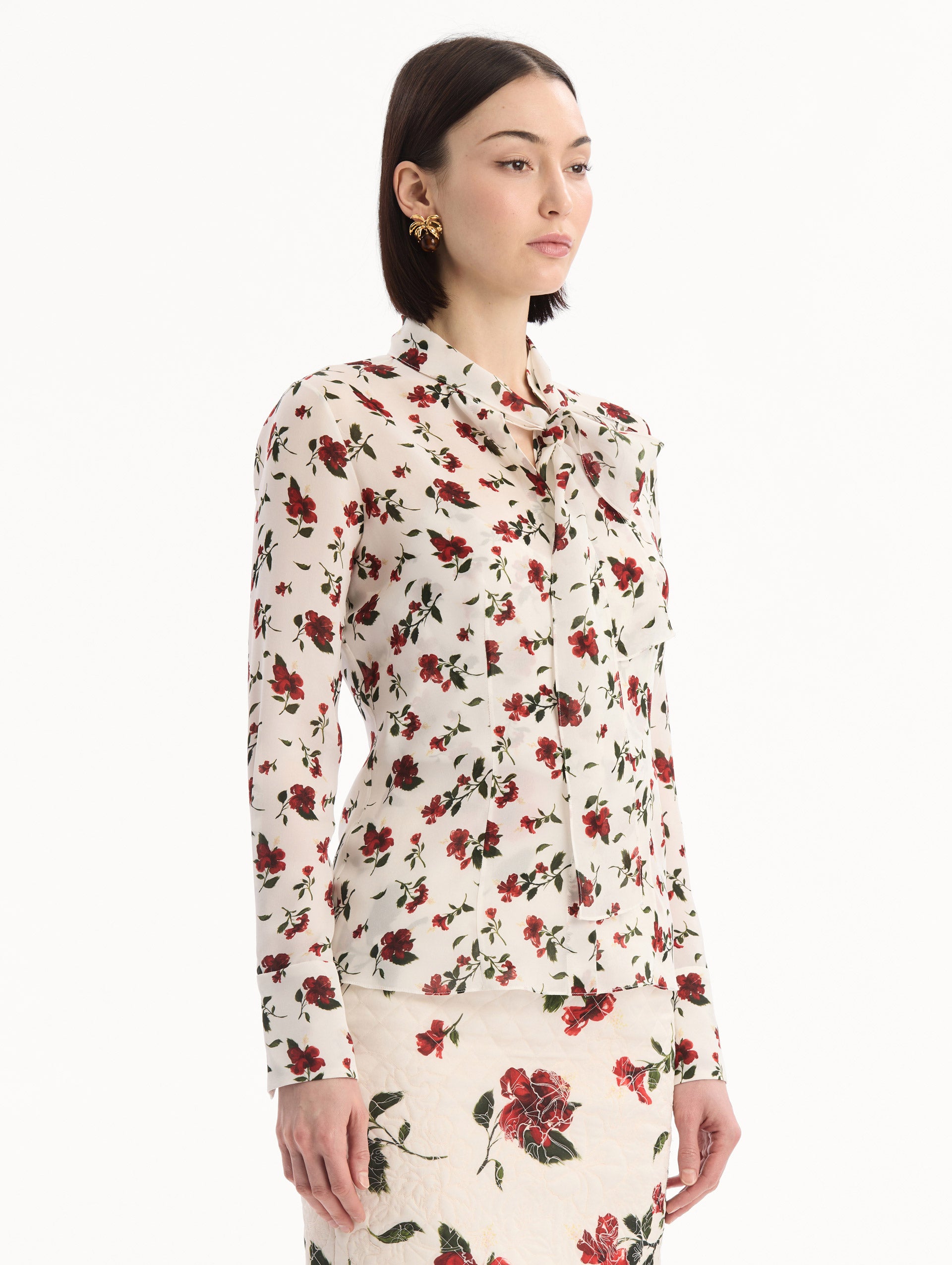 The model is wearing the Hibiscus Silk Chiffon Blouse in white printed with red hibiscus florals. Featuring a tie at neckline, long sleeves, and tailored silhouette. Side View.