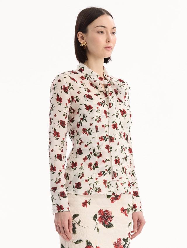 The model is wearing the Hibiscus Silk Chiffon Blouse in white printed with red hibiscus florals. Featuring a tie at neckline, long sleeves, and tailored silhouette. Side View.