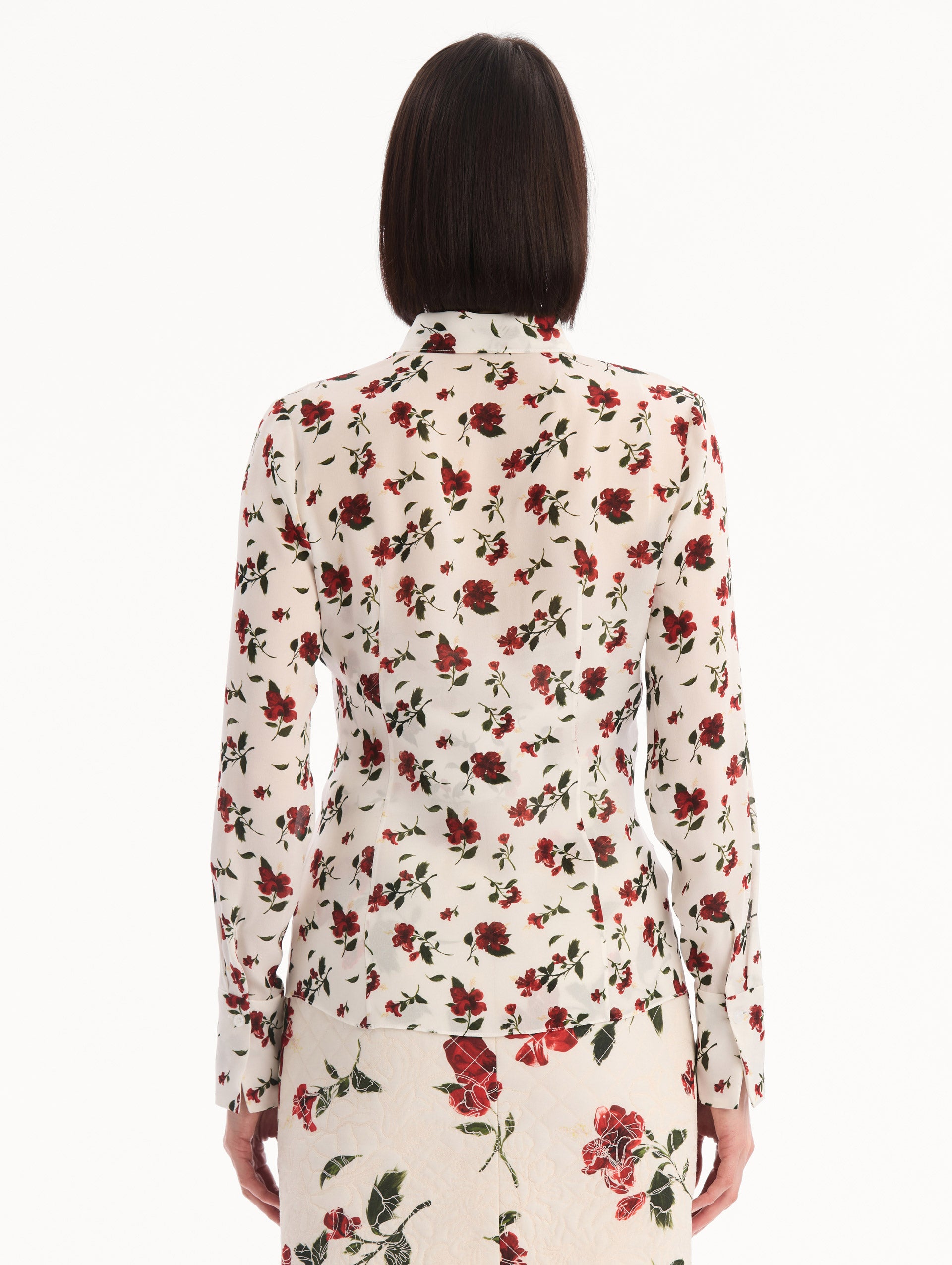 The model is wearing the Hibiscus Silk Chiffon Blouse in white printed with red hibiscus florals. Featuring a tie at neckline, long sleeves, and tailored silhouette. Back View.
