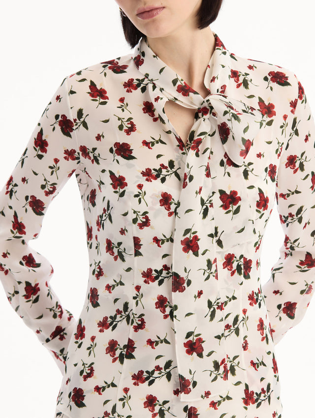 The model is wearing the Hibiscus Silk Chiffon Blouse in white printed with red hibiscus florals. Featuring a tie at neckline, long sleeves, and tailored silhouette. Detail View.