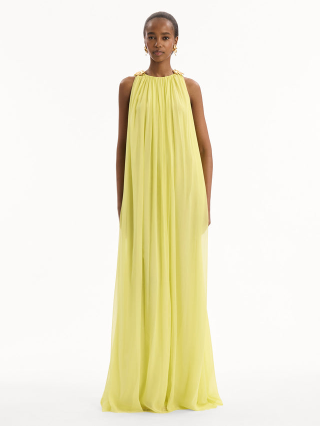 The model is wearing the Hibiscus-Detail Silk Chiffon Caftan in a light yellow-green color. Featuring a draping, gathered silhouette, long skirt, and sculptural hibiscus details at the shoulders. Front View.