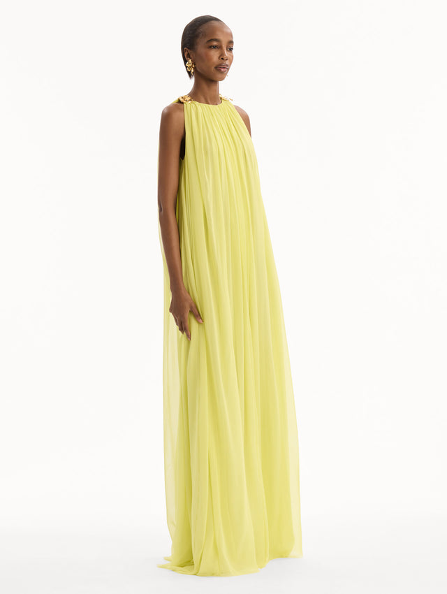 The model is wearing the Hibiscus-Detail Silk Chiffon Caftan in a light yellow-green color. Featuring a draping, gathered silhouette, long skirt, and sculptural hibiscus details at the shoulders. Side View.