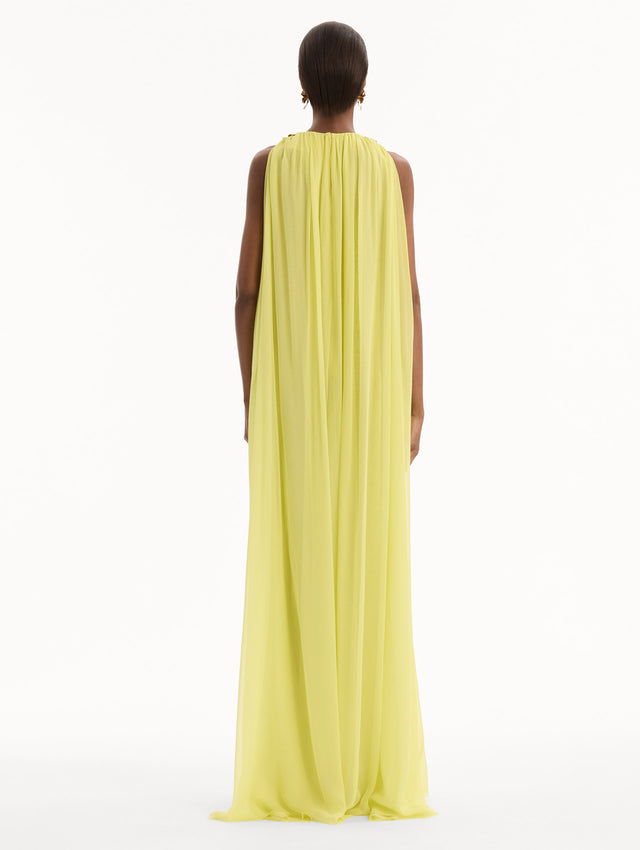 The model is wearing the Hibiscus-Detail Silk Chiffon Caftan in a light yellow-green color. Featuring a draping, gathered silhouette, long skirt, and sculptural hibiscus details at the shoulders. Back View.
