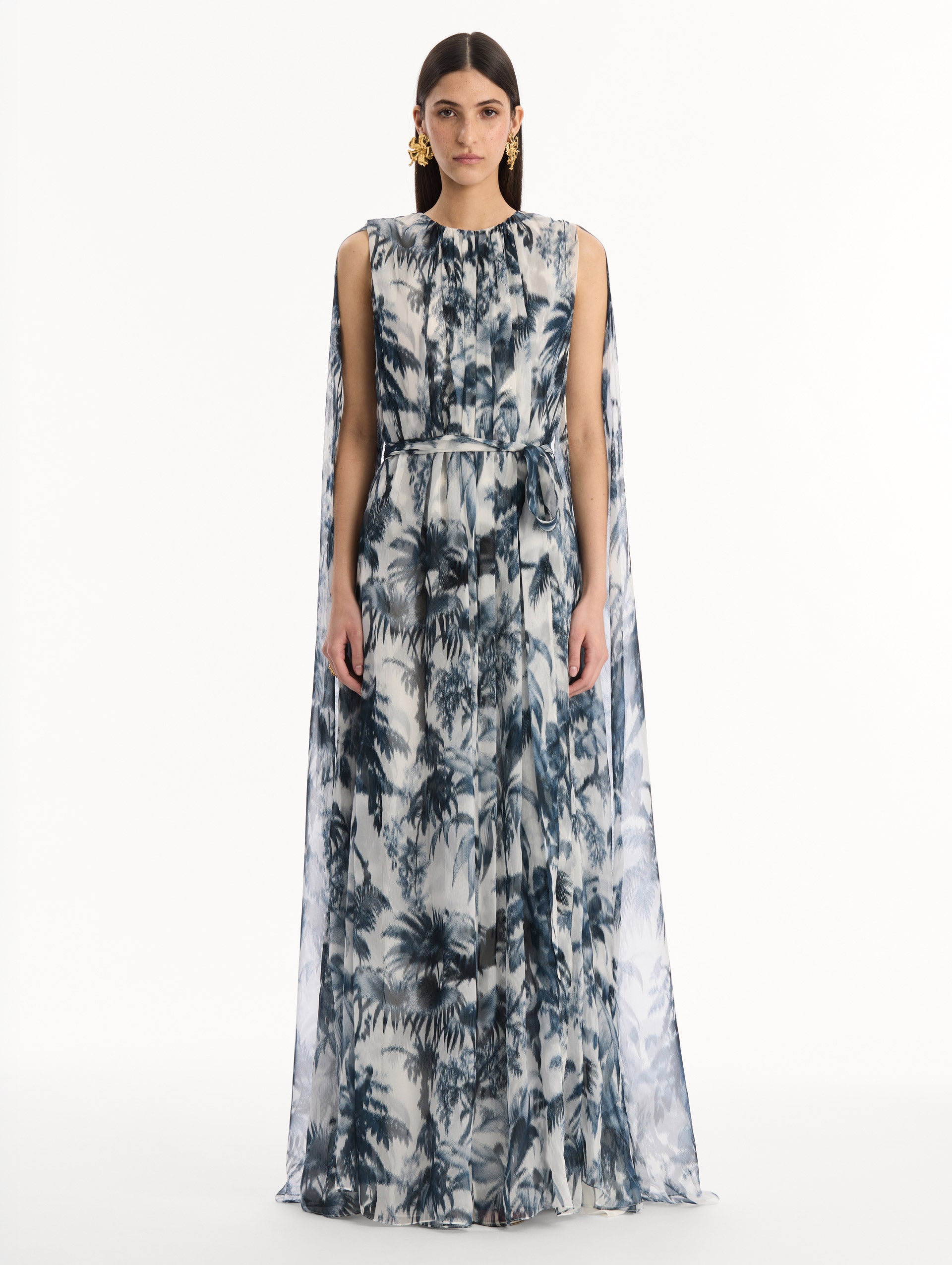 The model is wearing the Chiné Palm Leaves Silk Caftan White with Navy printed palm leaves. Featuring a gathered neckline, cape at back, flowing silhouette, and tie at waist. Front View.