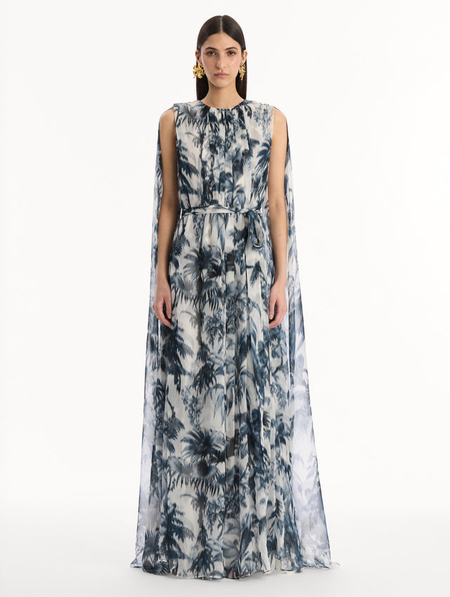 The model is wearing the Chiné Palm Leaves Silk Caftan White with Navy printed palm leaves. Featuring a gathered neckline, cape at back, flowing silhouette, and tie at waist. Front View.