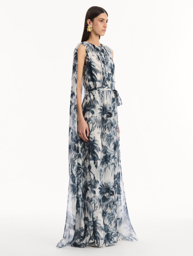 The model is wearing the Chiné Palm Leaves Silk Caftan White with Navy printed palm leaves. Featuring a gathered neckline, cape at back, flowing silhouette, and tie at waist. Side View.