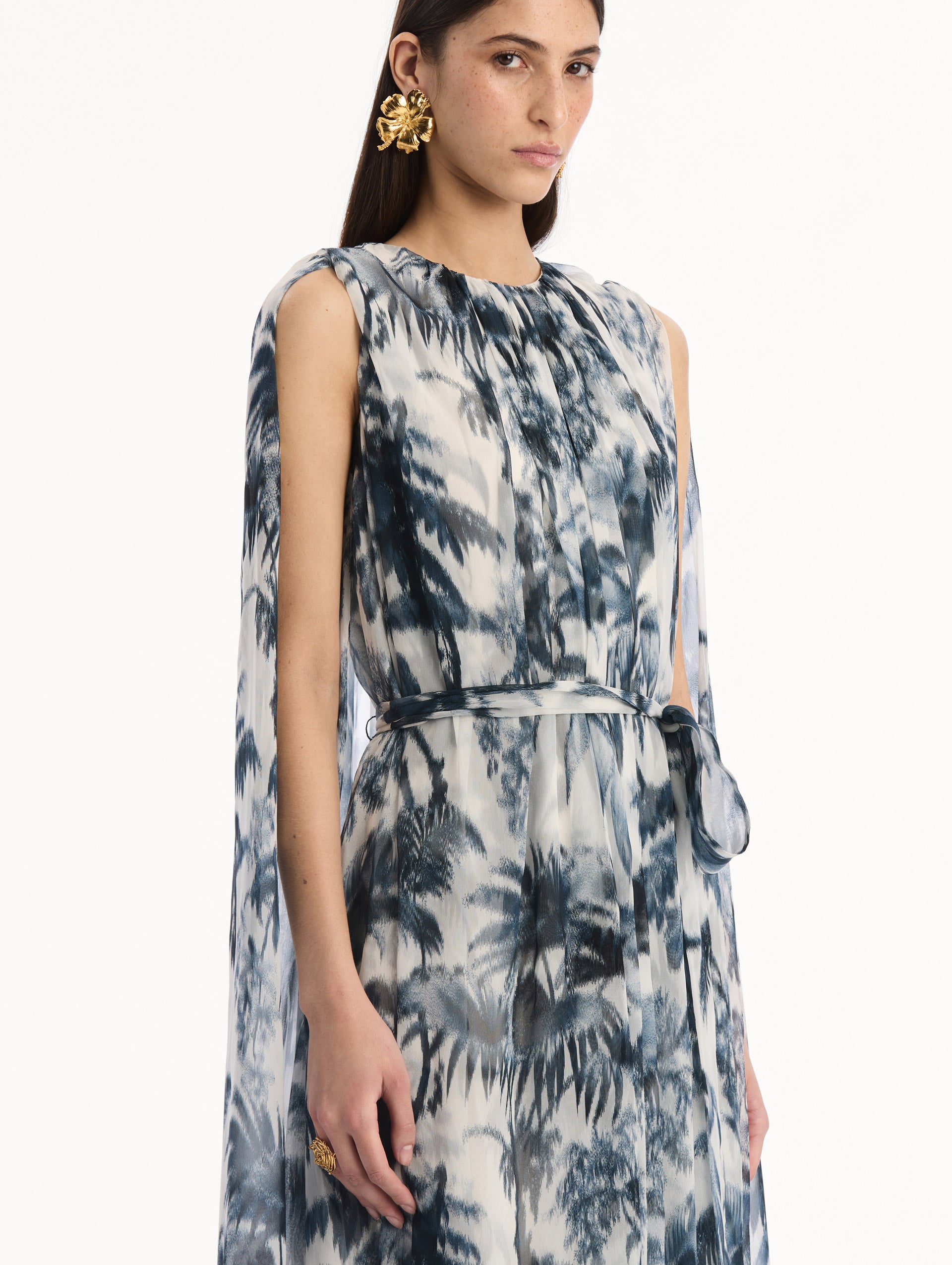 The model is wearing the Chiné Palm Leaves Silk Caftan White with Navy printed palm leaves. Featuring a gathered neckline, cape at back, flowing silhouette, and tie at waist. Detail View.