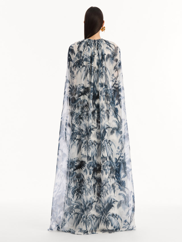 The model is wearing the Chiné Palm Leaves Silk Caftan White with Navy printed palm leaves. Featuring a gathered neckline, cape at back, flowing silhouette, and tie at waist. Back View.