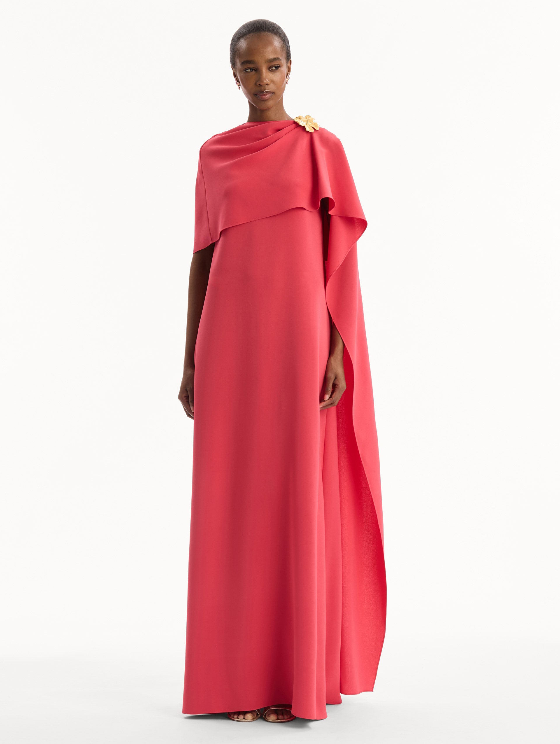 The model is wearing the Hibiscus Brooch Georgette Caftan in Hot Pink. Featuring a softly structured cape silhouette and a sculptural hibiscus brooch accent at the shoulder. Front View.