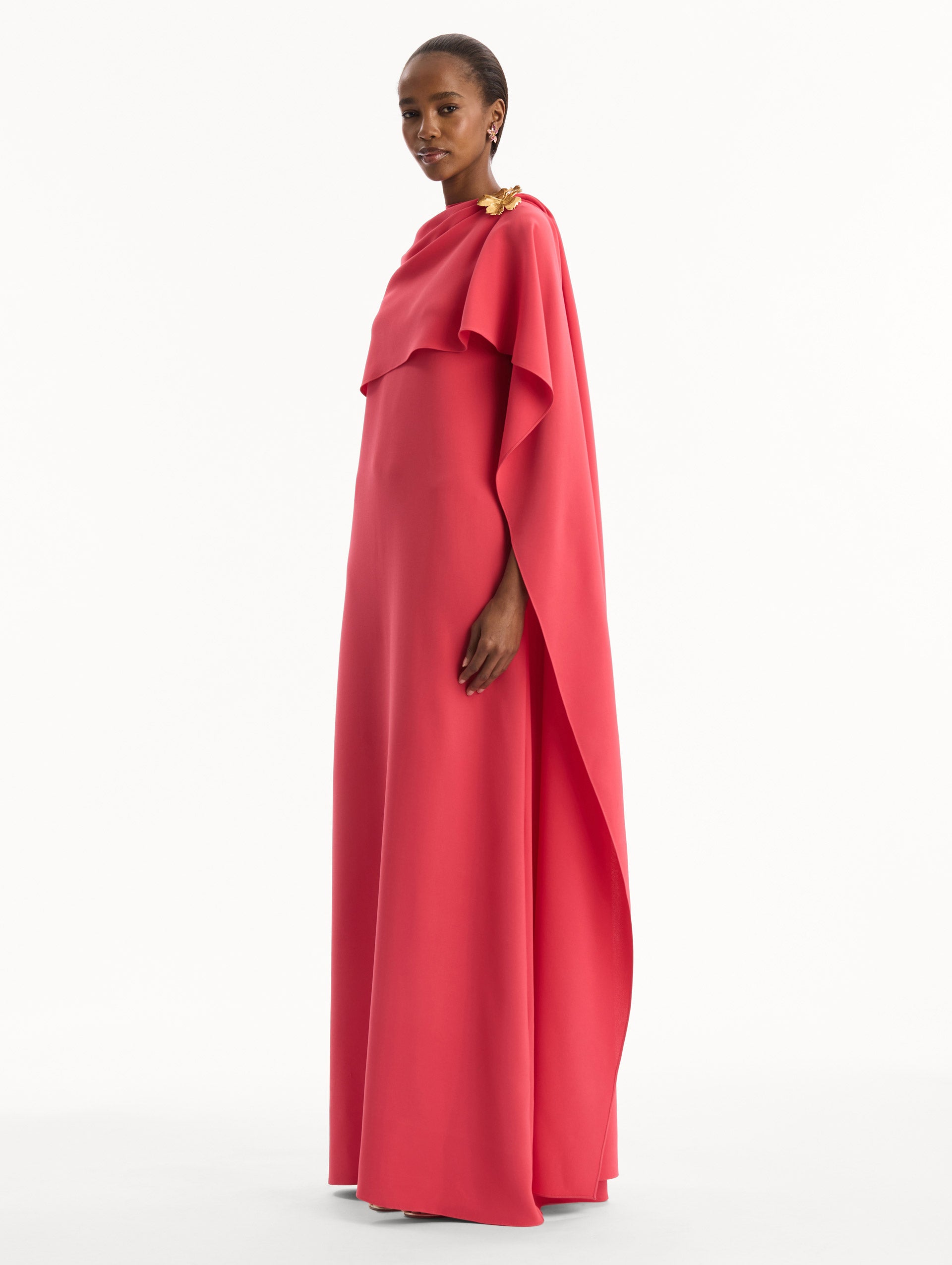 The model is wearing the Hibiscus Brooch Georgette Caftan in Hot Pink. Featuring a softly structured cape silhouette and a sculptural hibiscus brooch accent at the shoulder. Side View.