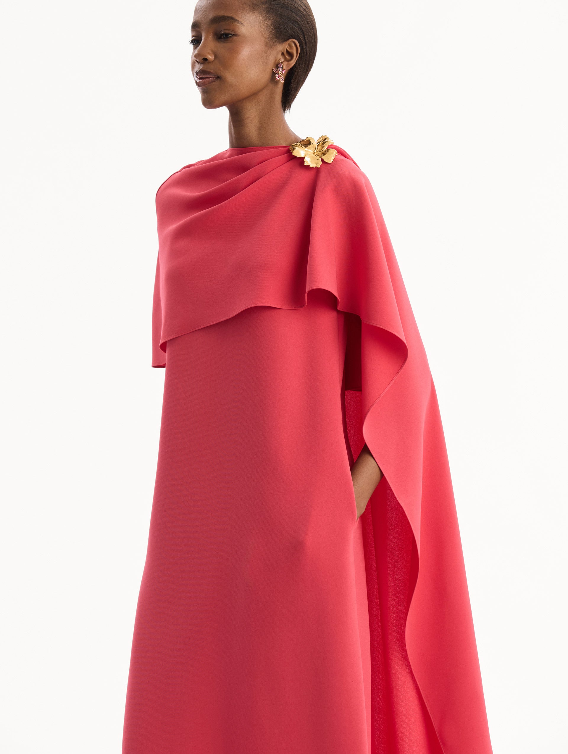 The model is wearing the Hibiscus Brooch Georgette Caftan in Hot Pink. Featuring a softly structured cape silhouette and a sculptural hibiscus brooch accent at the shoulder. Detail View.