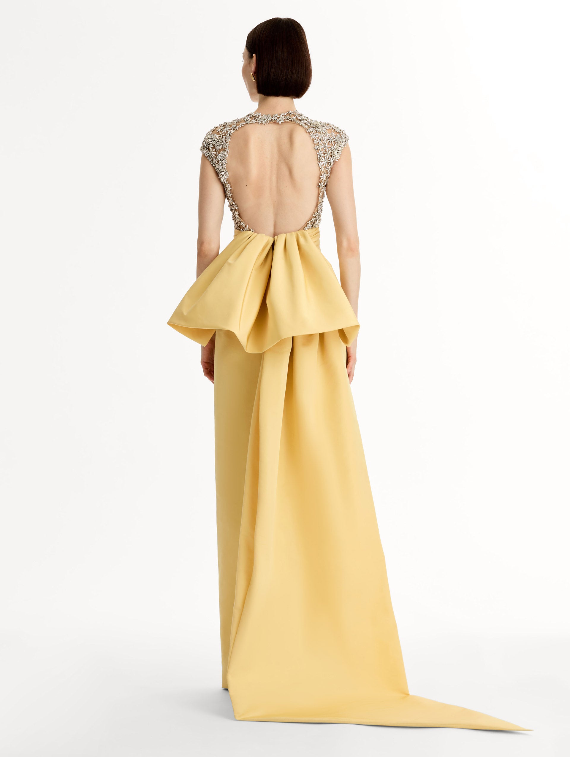 Yellow floor length gown with crystal porcelain flowers on an open back sculpted bodice. Dramatic faille skirt with cascading peplum train. Back View.