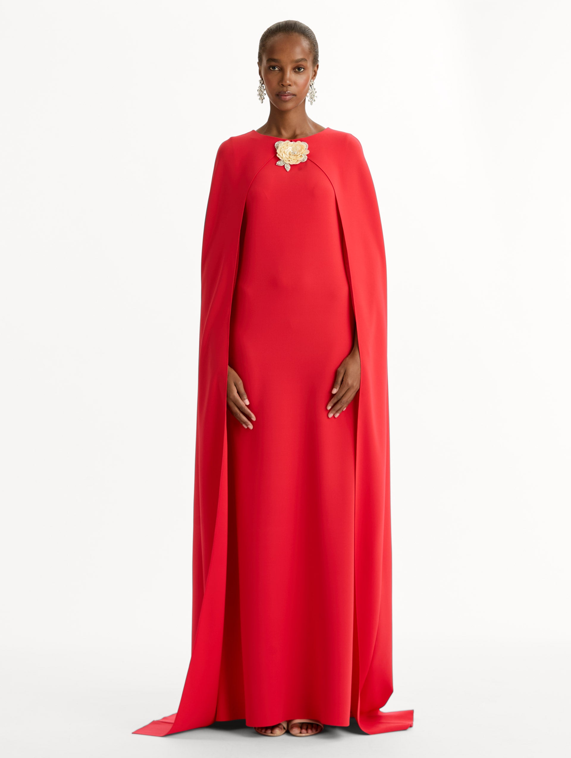 The model is wearing the Brooch Embroidered Cape-Back Caftan in the color red featuring draped sleeves and a floral brooch at center neckline. Front View.