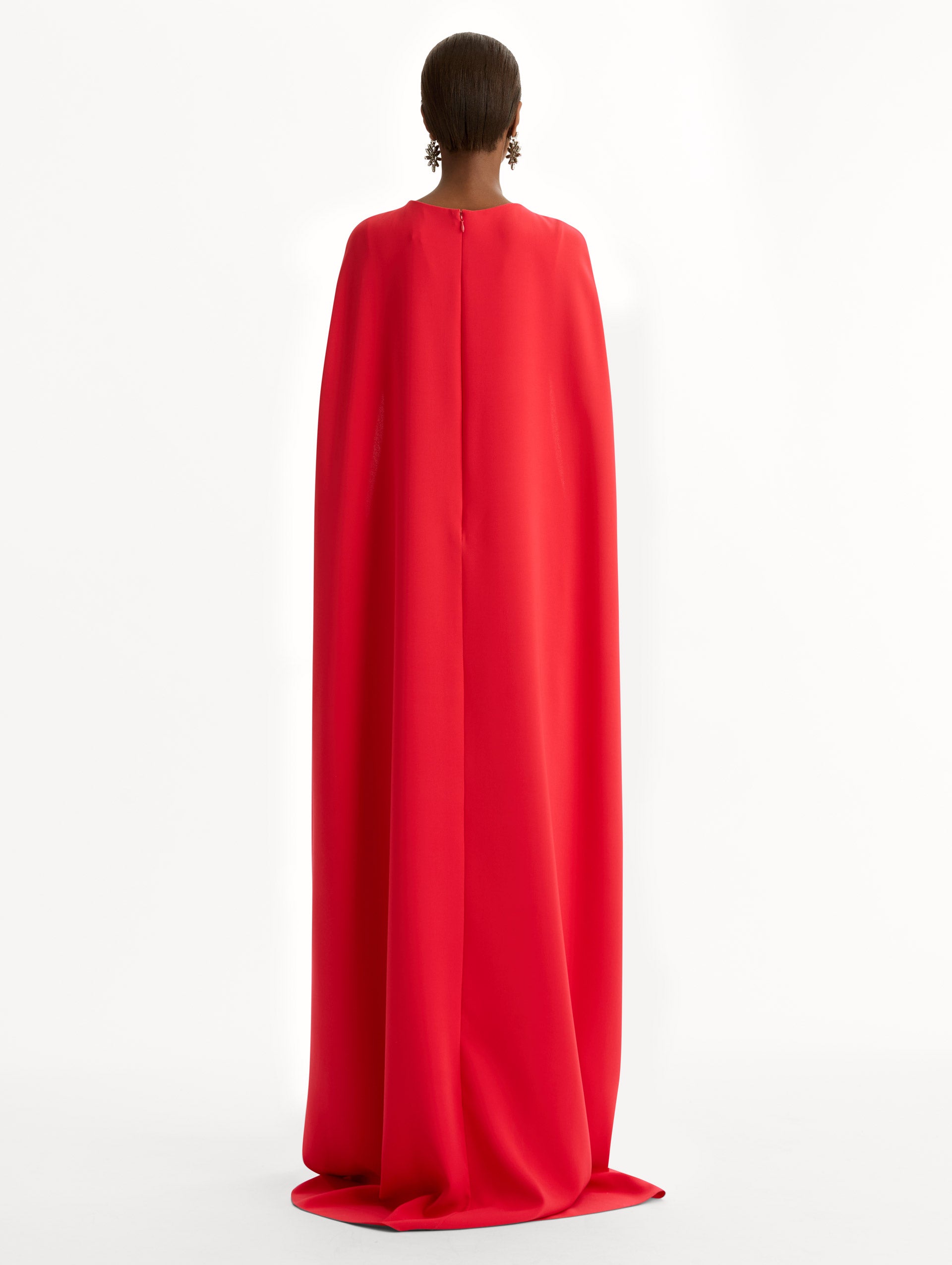 The model is wearing the Brooch Embroidered Cape-Back Caftan in the color red featuring draped sleeves and a floral brooch at center neckline. Back View.