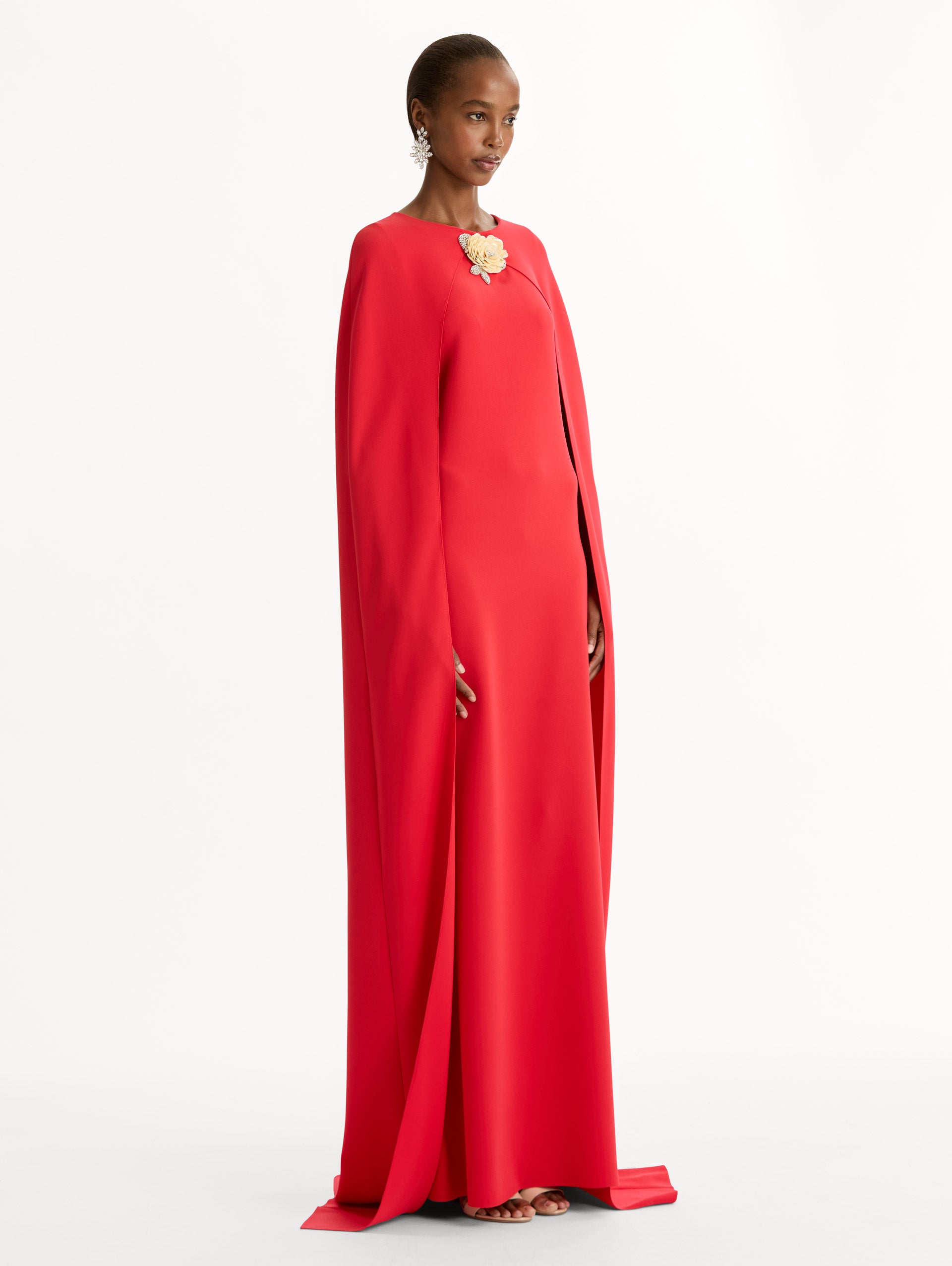 The model is wearing the Brooch Embroidered Cape-Back Caftan in the color red featuring draped sleeves and a floral brooch at center neckline. Side View.