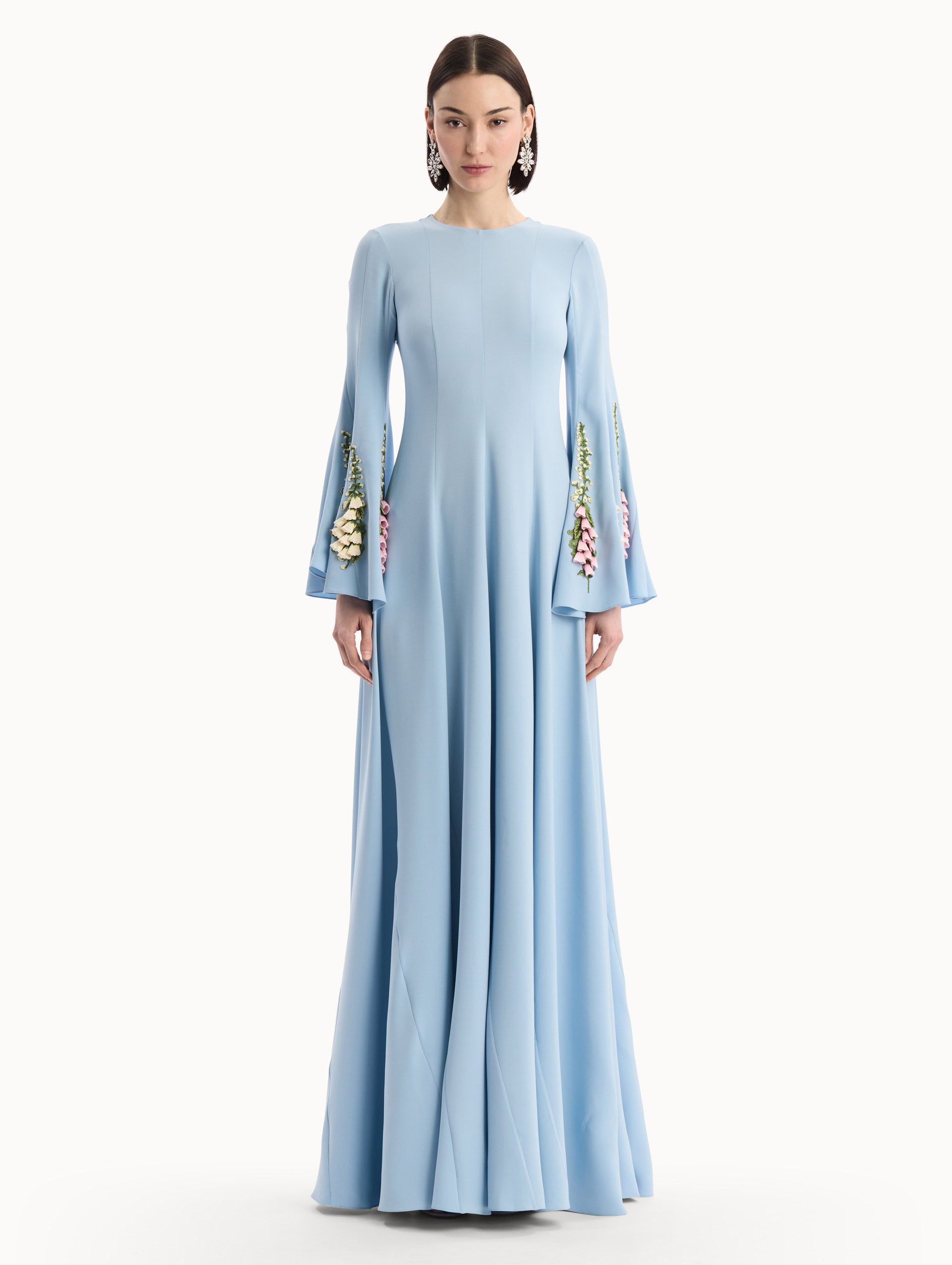 The model is wearing the Foxglove Embroidered Bell Sleeve Caftan in light blue featuring a floor-sweeping hemline and wide, bell sleeves embroidered with floral motifs. Front View.