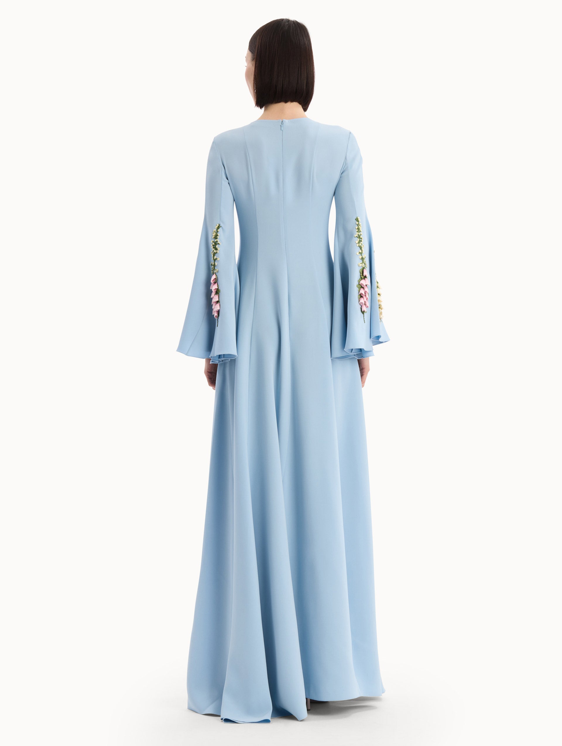 The model is wearing the Foxglove Embroidered Bell Sleeve Caftan in light blue featuring a floor-sweeping hemline and wide, bell sleeves embroidered with floral motifs. Back View.